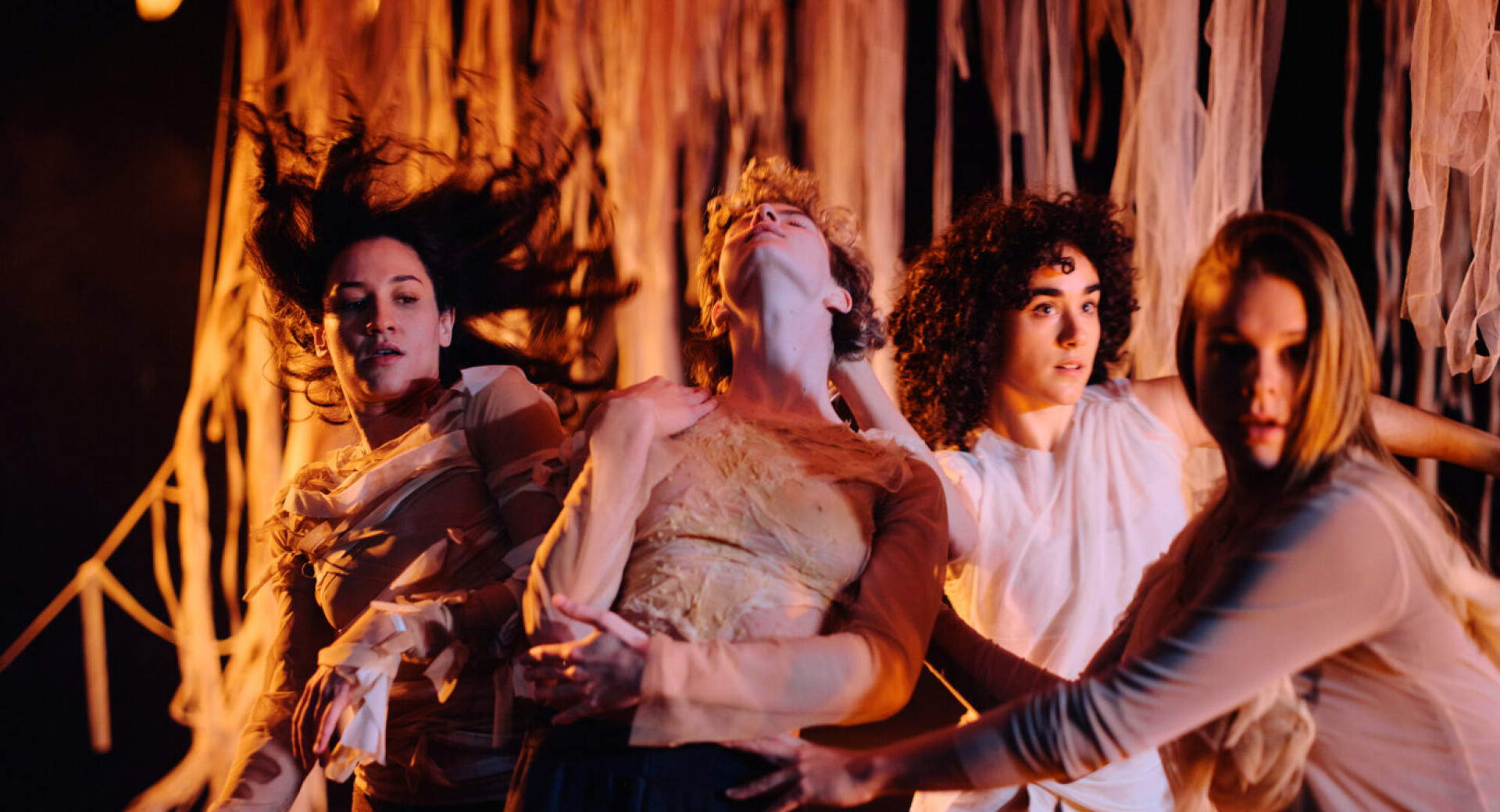 Stirje plesalci nastopajo na odru v oblacilih kozne barve, za njimi pa visijo prosojne zavese v kozni barvi.