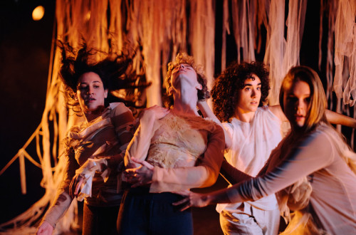 Stirje plesalci nastopajo na odru v oblacilih kozne barve, za njimi pa visijo prosojne zavese v kozni barvi.