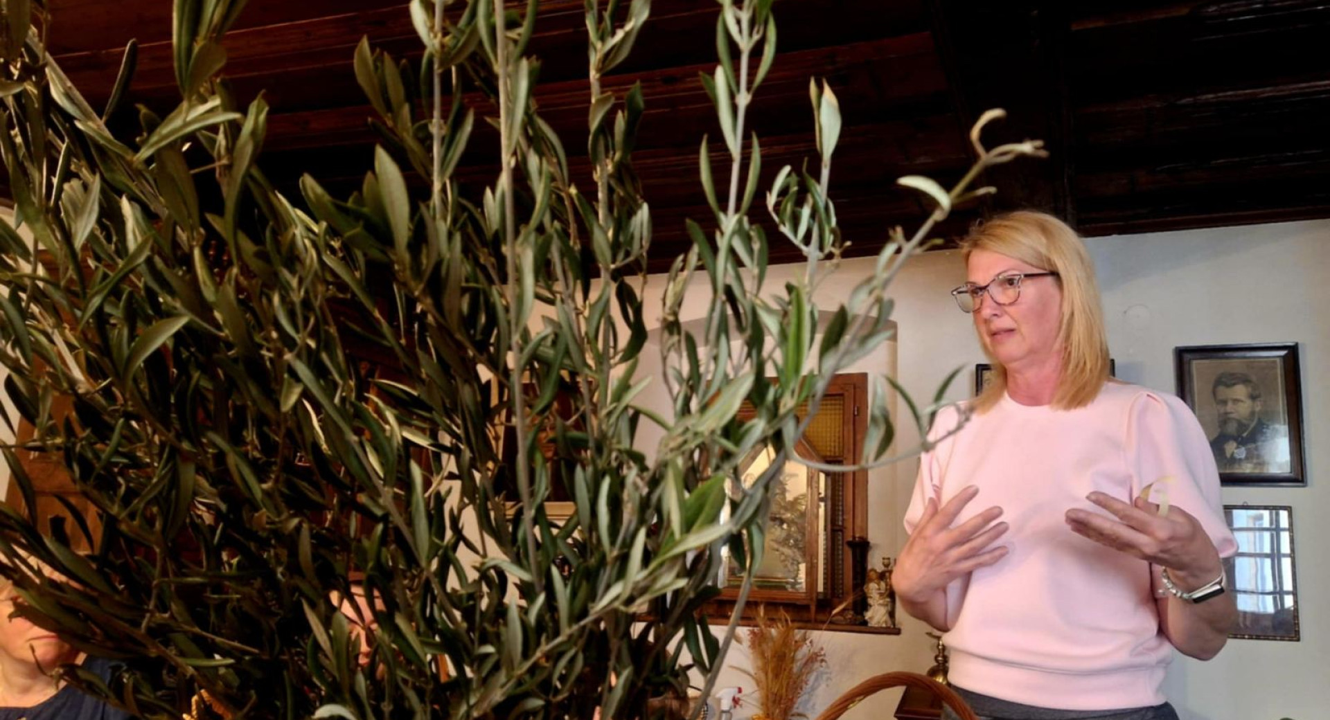 Plavolasa zenska z ocali v roznatem puloverju stoji poleg zelenja in nekaj razlaga.