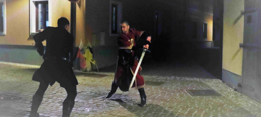 Two men in medieval clothing are fighting with swords on a street at dusk.