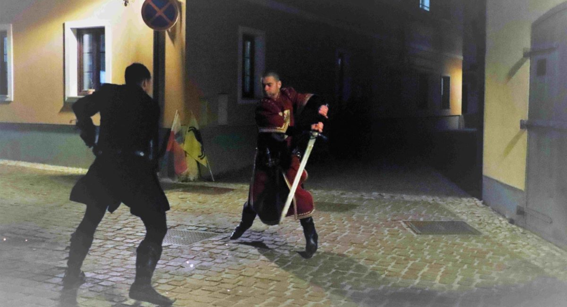 Two men in medieval clothing are fighting with swords on a street at dusk.
