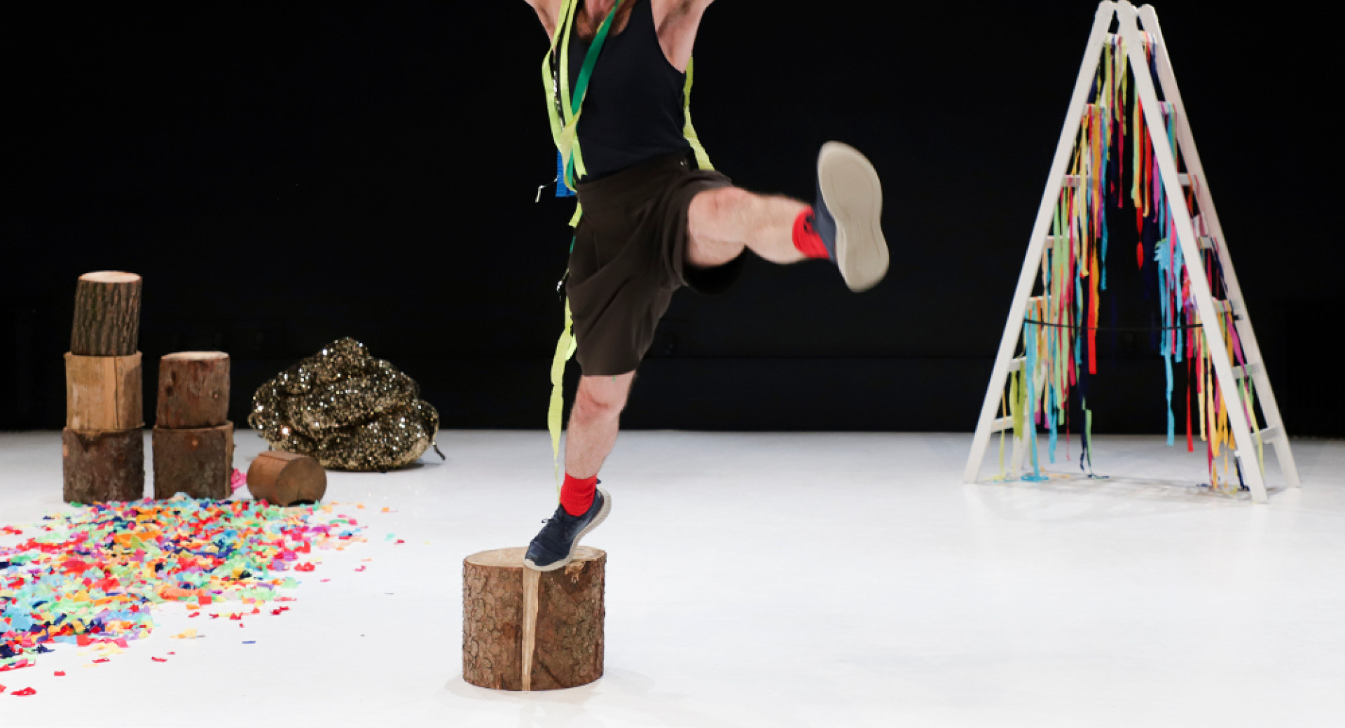A man is on a white stage with a black background; he is standing on a box with his right foot and jumping into the air, his left leg extended to the side and his arms raised upwards.