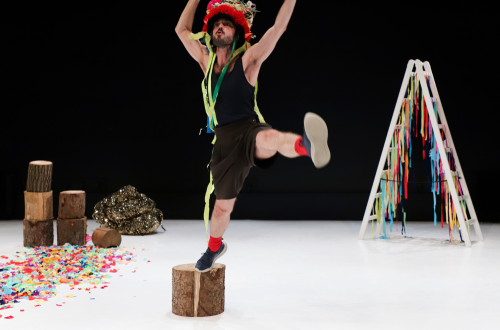 A man is on a white stage with a black background; he is standing on a box with his right foot and jumping into the air, his left leg extended to the side and his arms raised upwards.