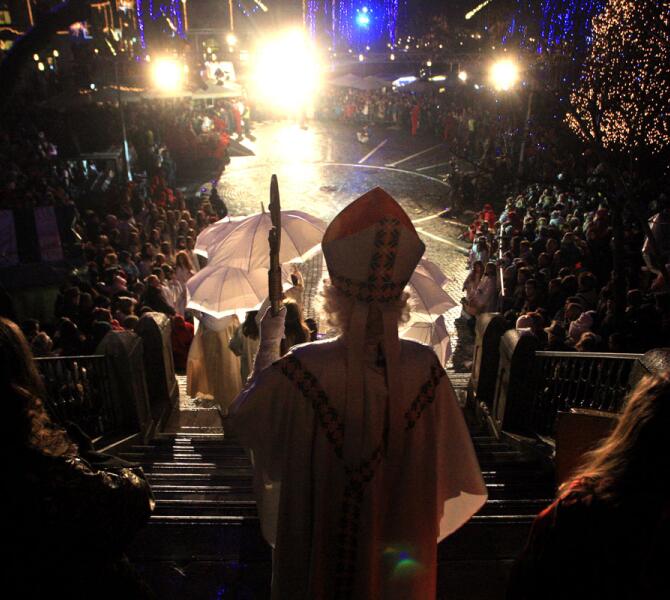 A figure dressed as St. Nicholas leads a festive procession at night, with a bright crowd and lights in the background.