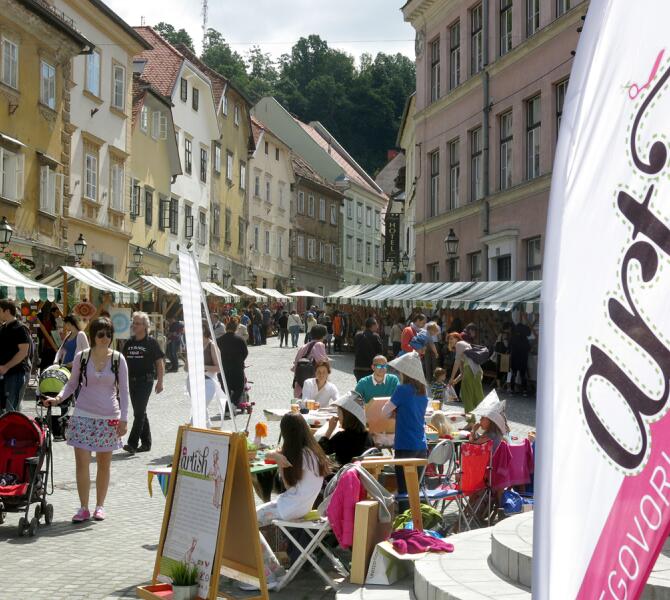 Živahna ulična tržnica v Ljubljani z ljudmi, ki se sprehajajo mimo stojnic, ter otroki, ki sodelujejo v dejavnostih za mizo.