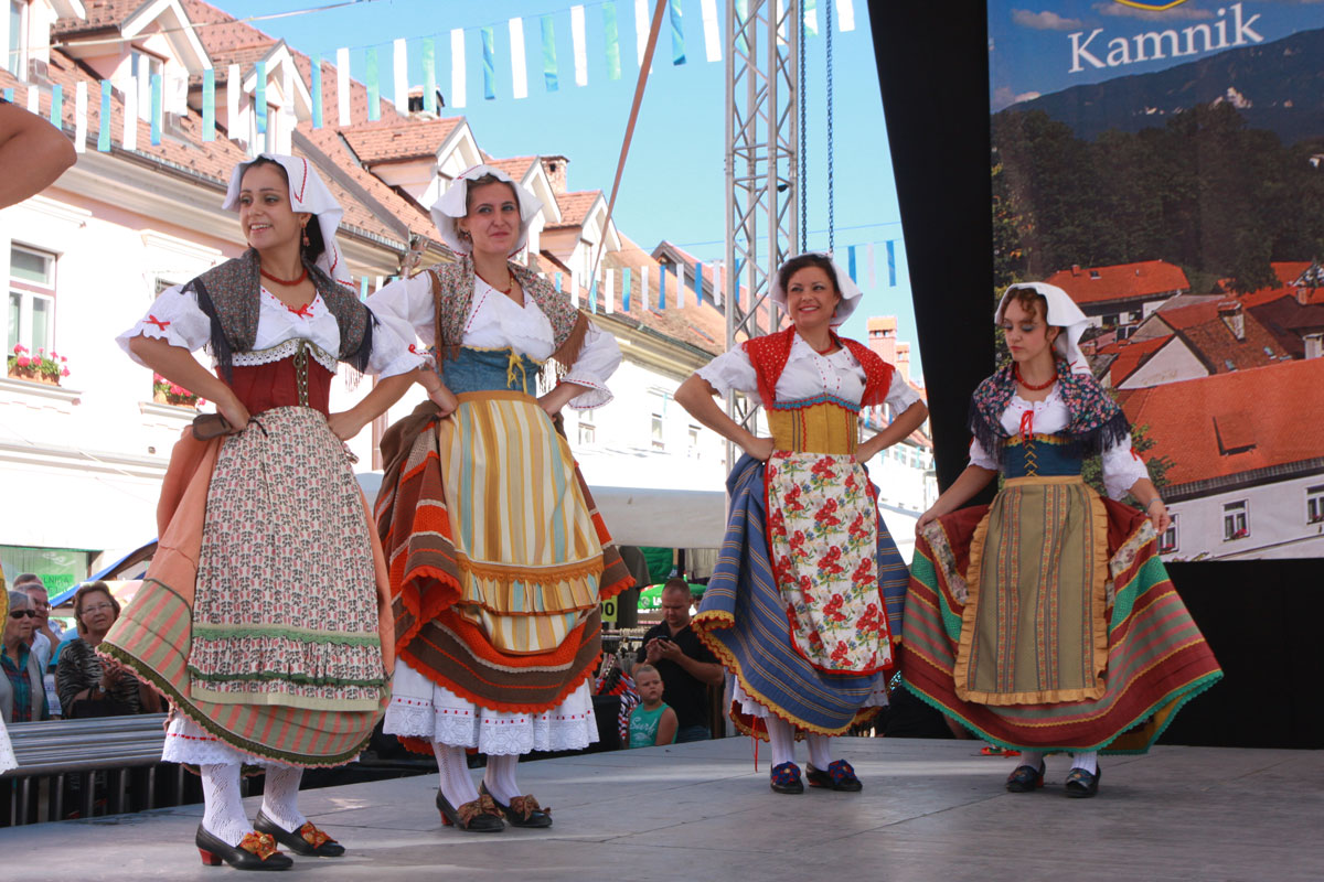 On stage, four women dressed in traditional Slovene costumes adorned with colorful patterns are dancing.