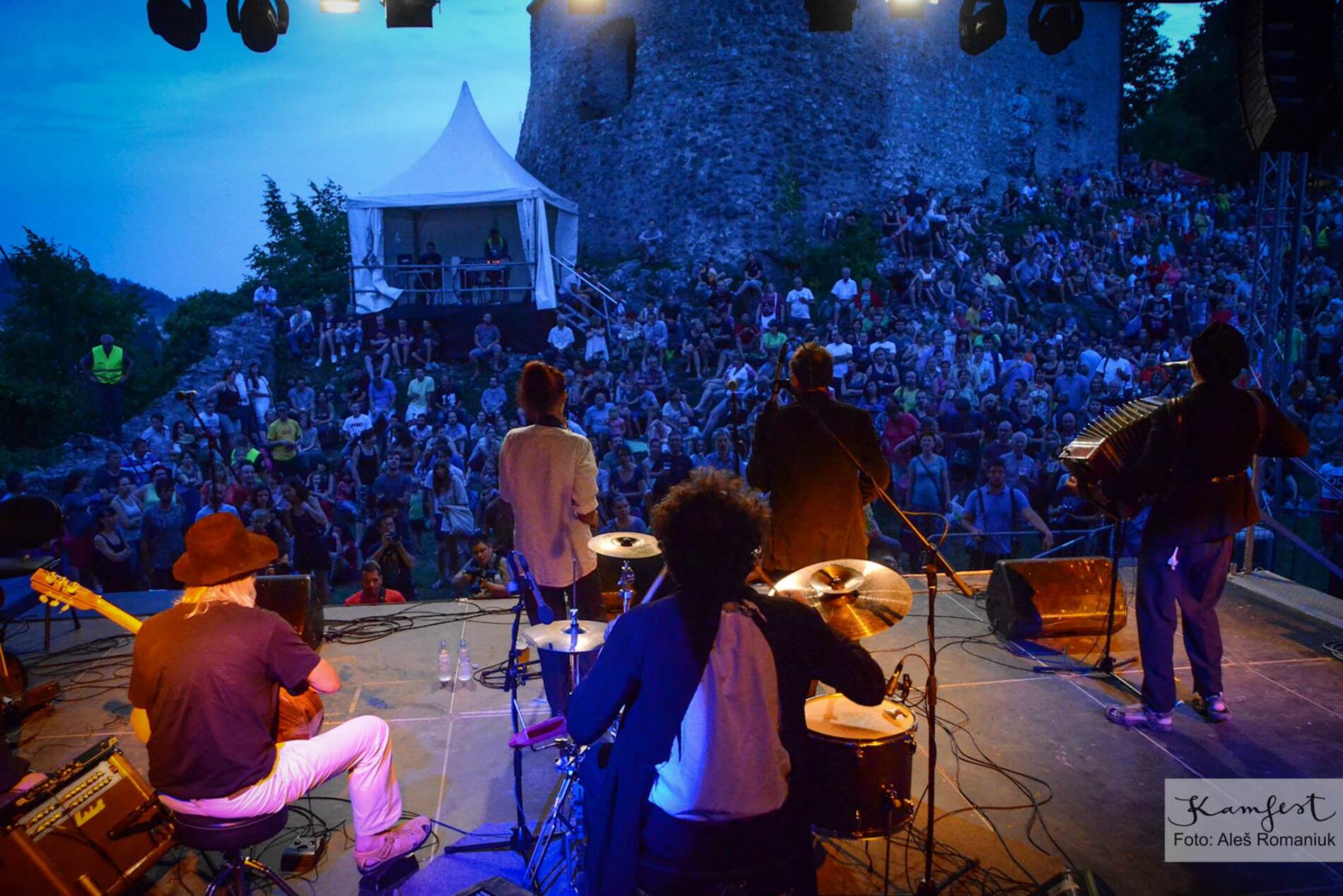 Musicians perform on stage in front of a large audience at dusk, with a historic stone building in the background.