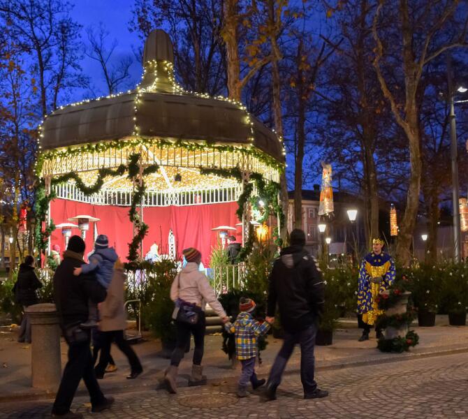 A festive gazebo decorated with lights and garlands in a park. Families and a man dressed in a festive costume are visible.