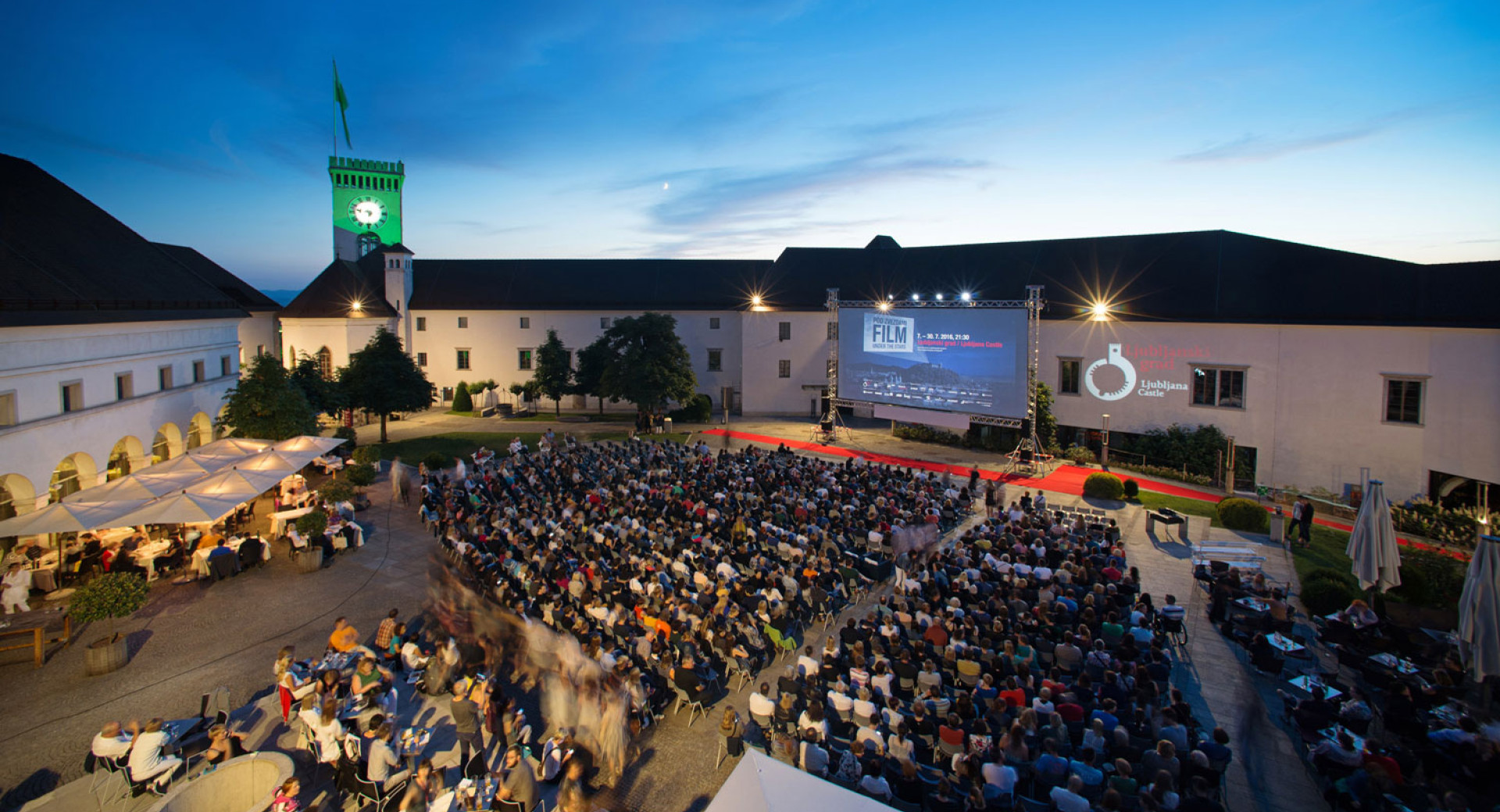 Na notranjem dvoriscu Ljubljanskega gradu obiskovalci sedijo na stolih pred velikim platnom in si ogledujejo film; v ozadju se dviga Grajski stolp.