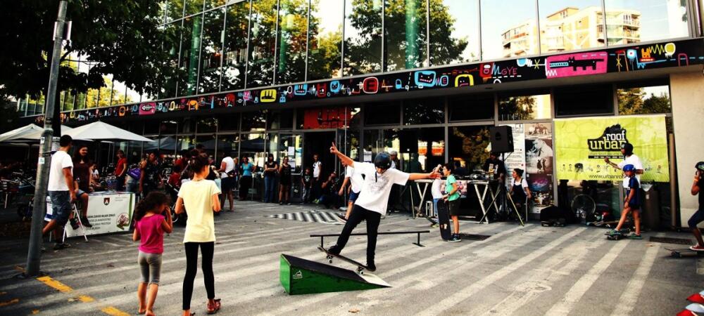 Des gens se rassemblent devant un grand bâtiment en verre pendant qu'un garçon fait du skateboard sur des obstacles devant lui.