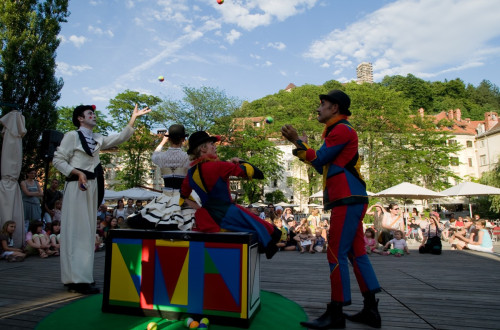 Two clowns and two jugglers are performing on stage, watched by the audience, with Castle Hill and Ljubljana Castle rising in the background.