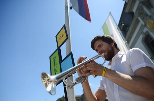 Moski, fotografiran od spodaj, igra na instrument, za njim pa sta zastava in modro nebo.