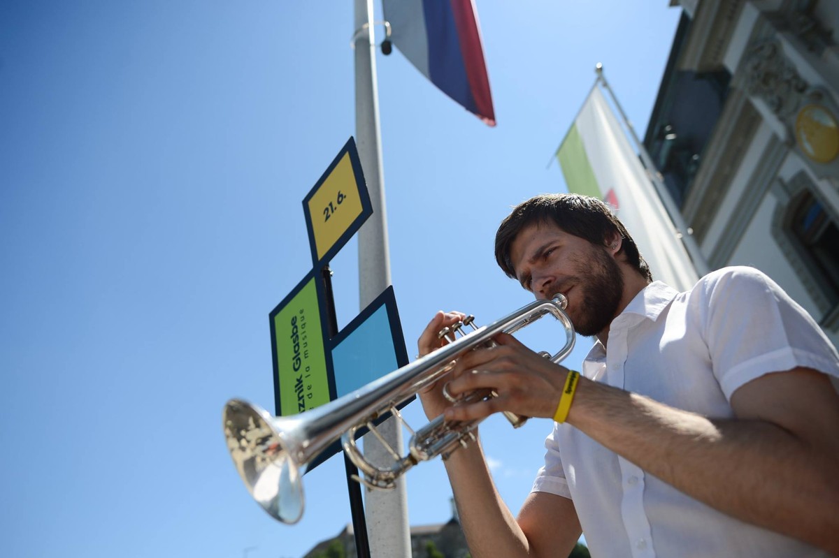 A man, photographed from a low angle, is playing an instrument with a flag and blue sky behind him.