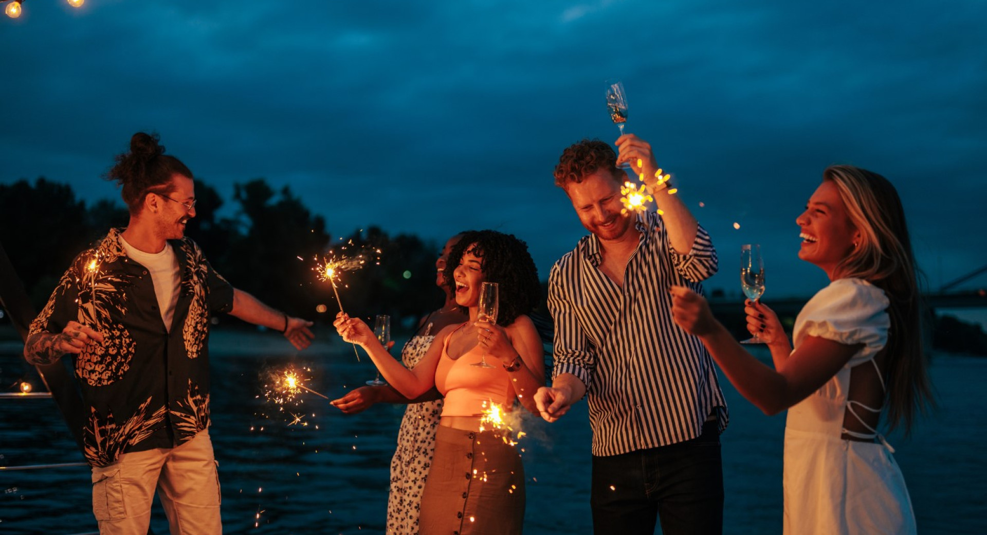Skupina mladih ljudi praznuje ob reki v vecernem casu, s sampanjcem in kresnickami v rokah, obdani z luckami in prijetno atmosfero.