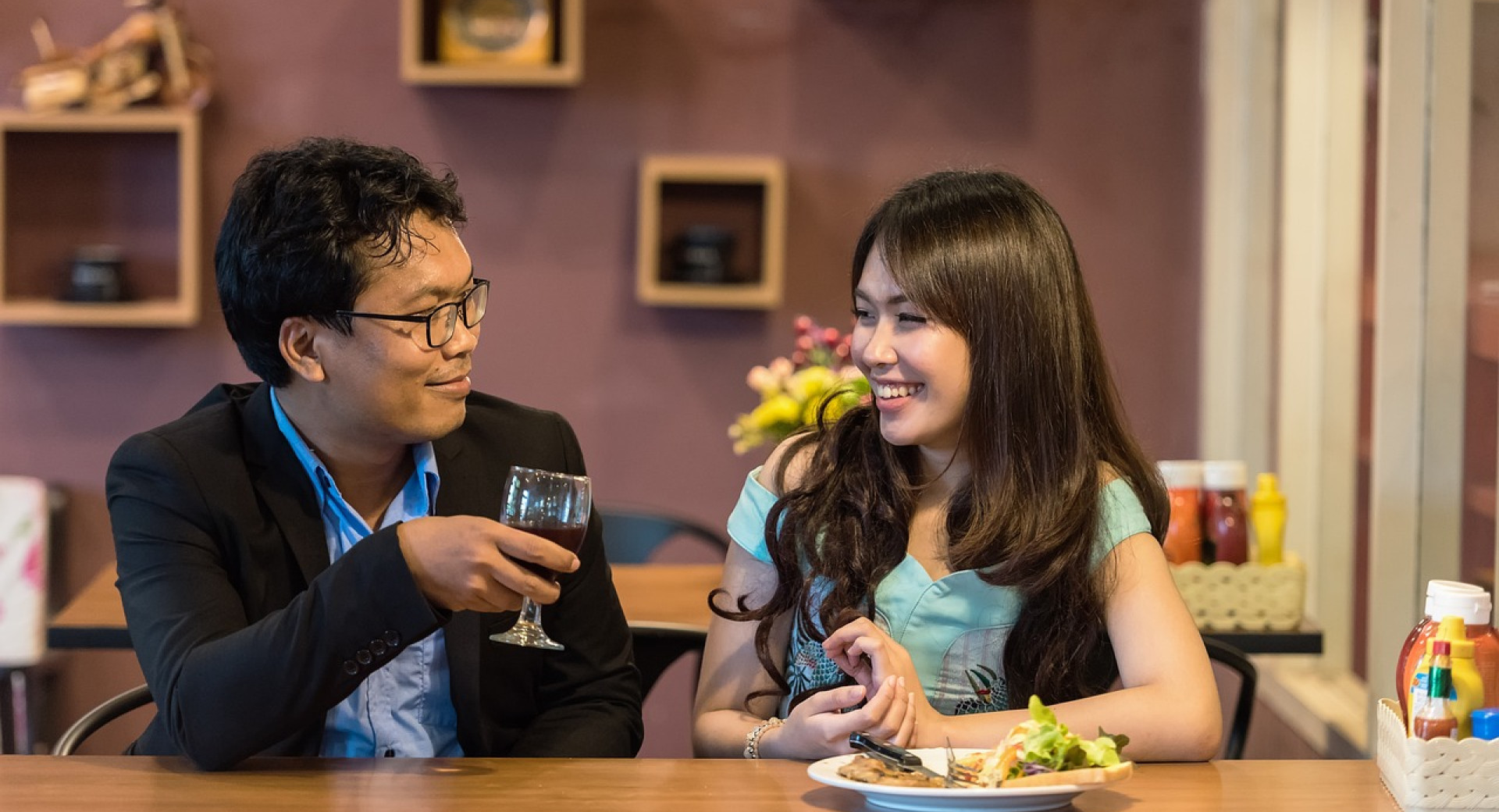 A man and a woman sit in a cosy café setting, smiling as they enjoy drinks and food on the table.