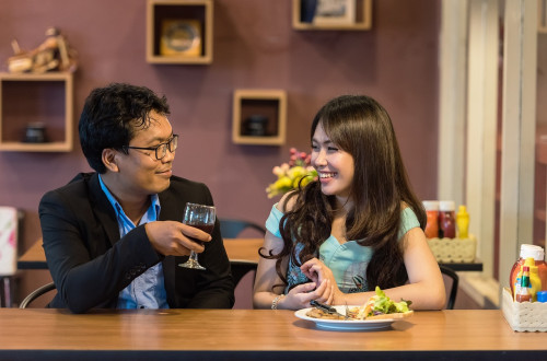 A man and a woman sit in a cosy café setting, smiling as they enjoy drinks and food on the table.