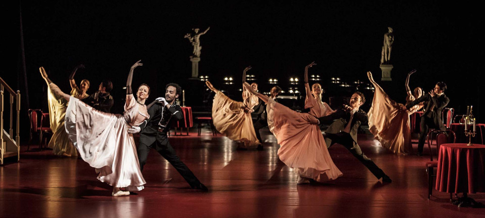 Skupina moskih in zenskih plesalcev v elegantnih oblekah med plesom na odru.