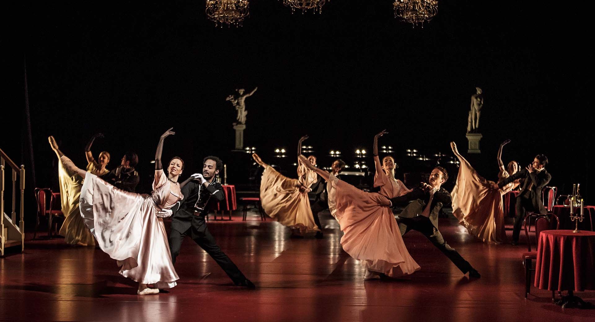 Skupina moskih in zenskih plesalcev v elegantnih oblekah med plesom na odru.