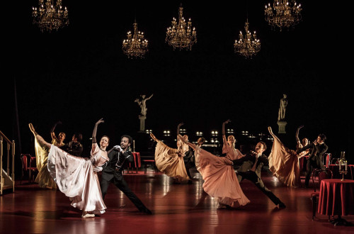 Skupina moskih in zenskih plesalcev v elegantnih oblekah med plesom na odru.