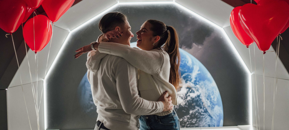 A young man and woman hugging and laughing together in a brightly lit, modern setting that resembles the interior of a space station