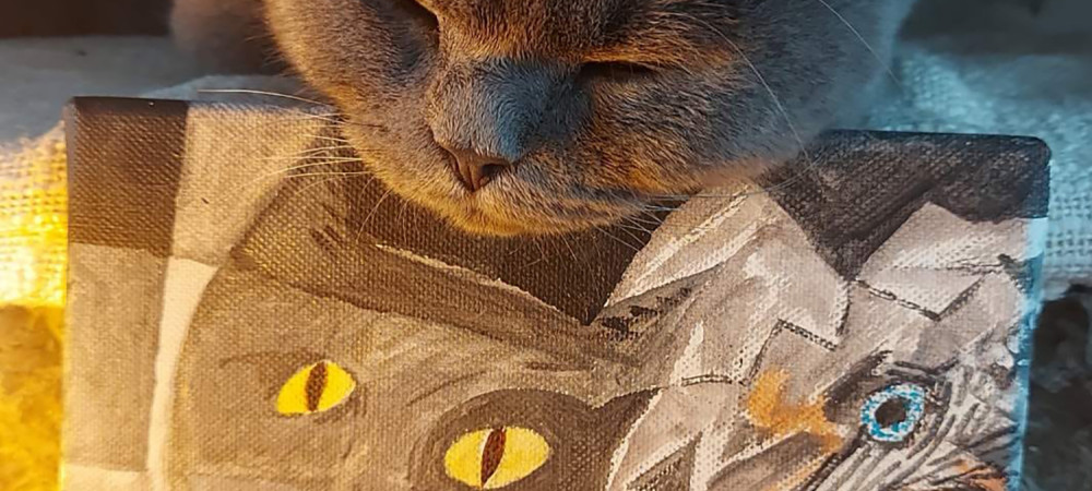 Siv macek z zaprtimi ocmi pociva svojo glavo na majhnem platnu, na katerem je naslikan portret macke v kubističnem slogu z izrazitimi rumenimi ocmi.