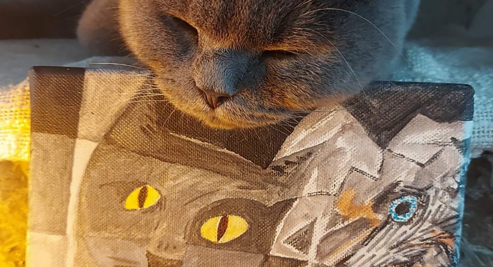 Siv macek z zaprtimi ocmi pociva svojo glavo na majhnem platnu, na katerem je naslikan portret macke v kubističnem slogu z izrazitimi rumenimi ocmi.