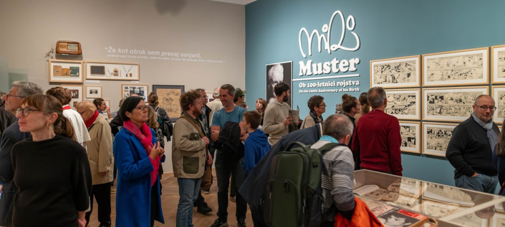 Ljudje na razstavi v turkizni sobi s stripovskimi ilustracijami na stenah.