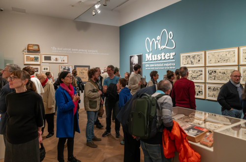 Ljudje na razstavi v turkizni sobi s stripovskimi ilustracijami na stenah.