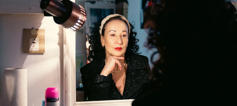 Zenska s skodranimi crnimi lasmi in rdeco sminko se gleda v ogledalo.