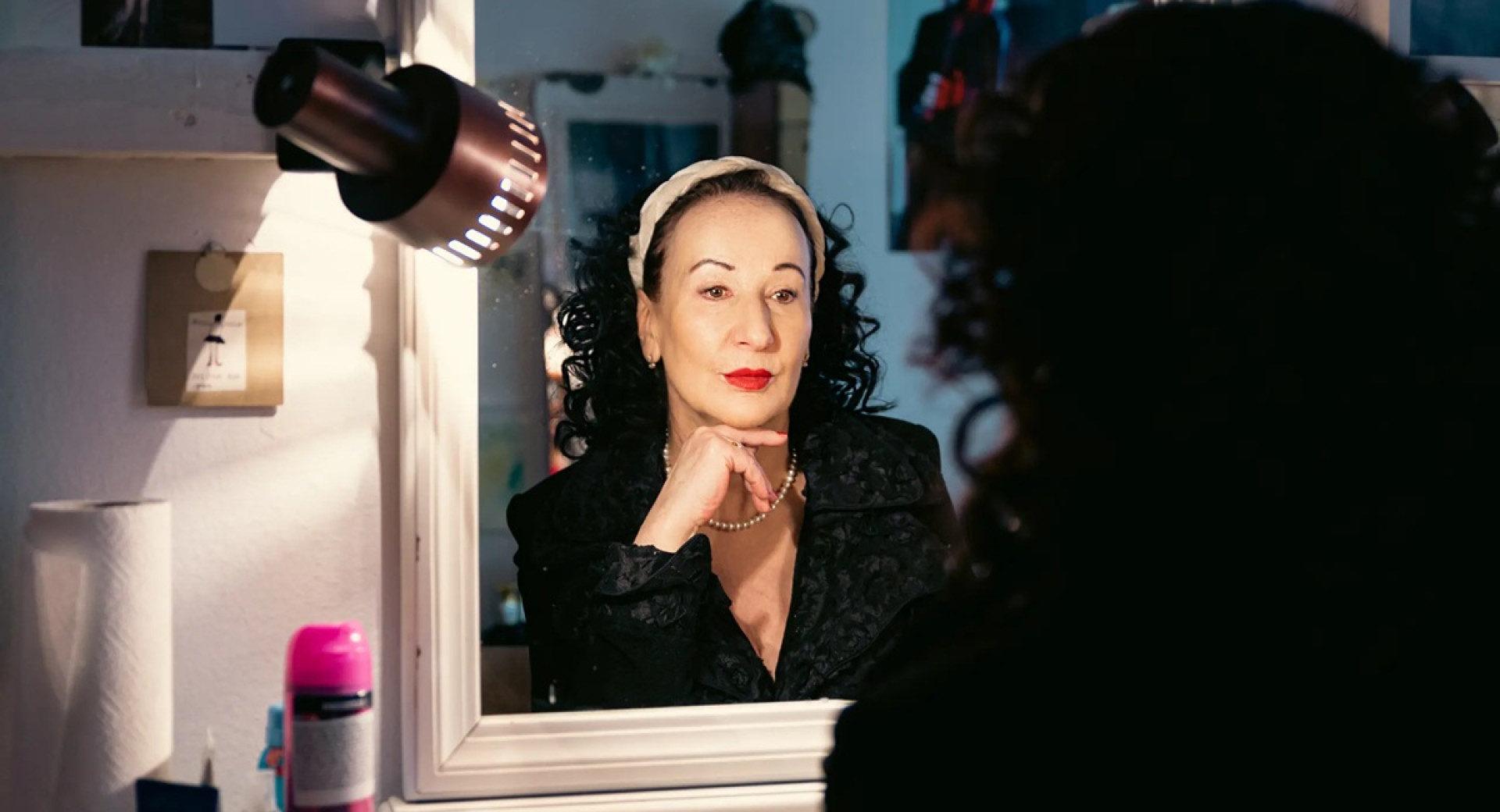 Zenska s skodranimi crnimi lasmi in rdeco sminko se gleda v ogledalo.