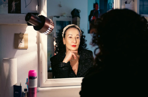 Zenska s skodranimi crnimi lasmi in rdeco sminko se gleda v ogledalo.