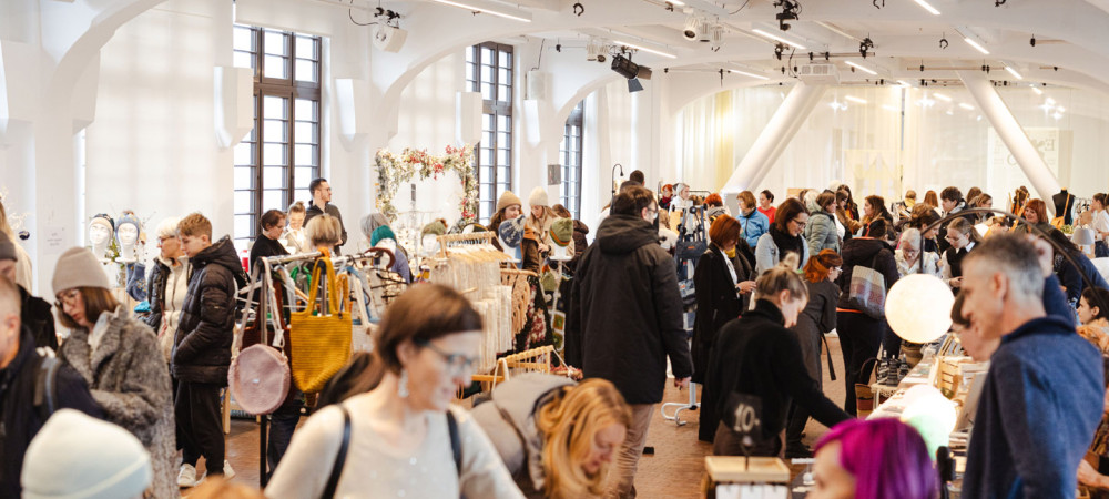 Živahen notranji umetniški sejem, poln ljudi, ki si ogledujejo različne stojnice.