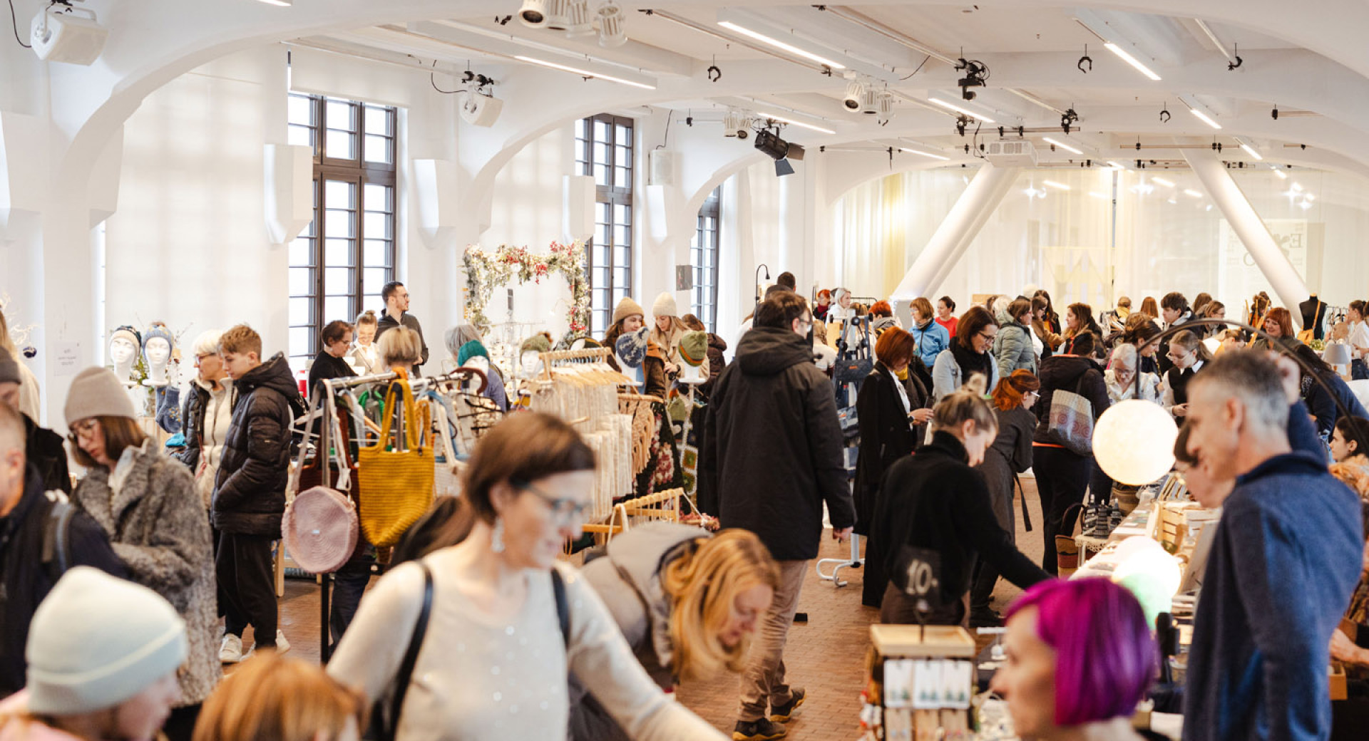 Živahen notranji umetniški sejem, poln ljudi, ki si ogledujejo različne stojnice.