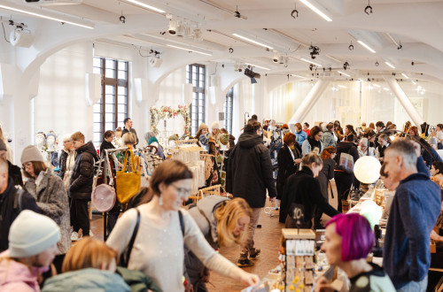 Živahen notranji umetniški sejem, poln ljudi, ki si ogledujejo različne stojnice.