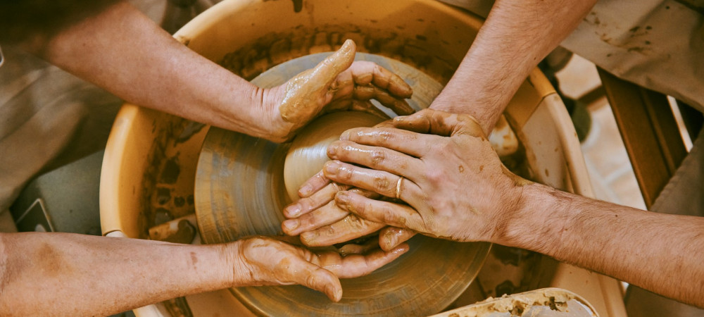 Bližnji posnetek iz visokega kota prikazuje štiri roke, prekrite z mokro, svetlo glino, ki skupaj delajo na lončarskem vretenu