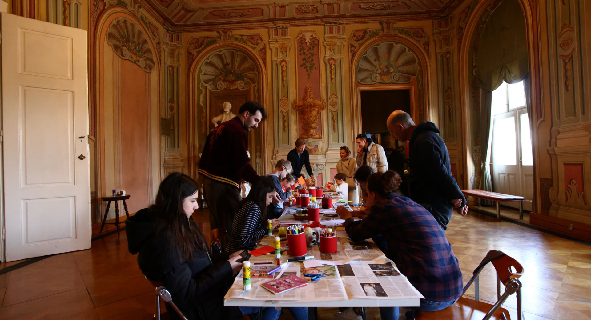 Fotografija ustvarjalne delavnice, ki poteka v dvorani z razkošnimi baročnimi freskami na stenah in stropu. Na sredini stoji dolga miza za katero sedi ali stoji več otrok in odraslih.