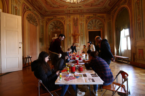 Fotografija ustvarjalne delavnice, ki poteka v dvorani z razkošnimi baročnimi freskami na stenah in stropu. Na sredini stoji dolga miza za katero sedi ali stoji več otrok in odraslih.