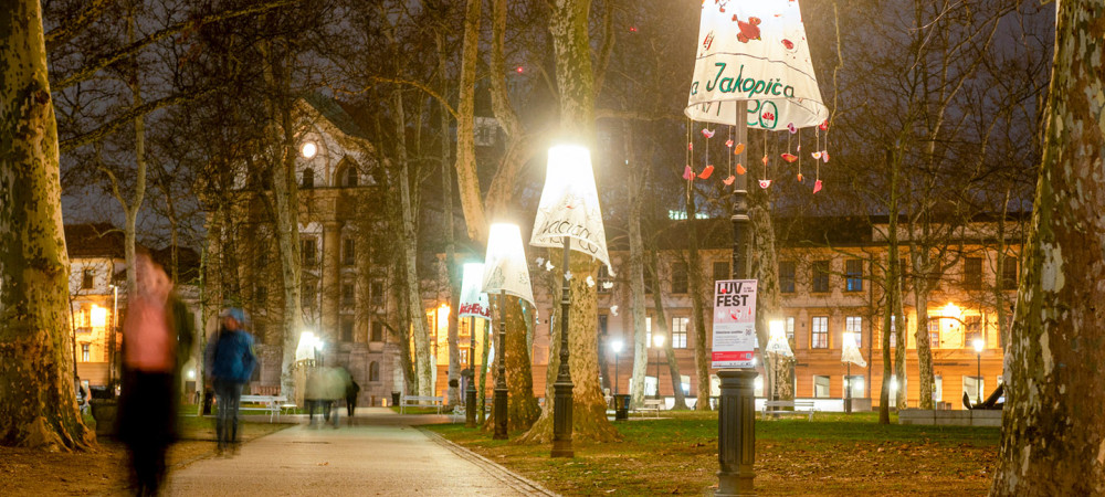 Fotografija tlakovane poti v parku ponoči, obdane z visokimi drevesi in uličnimi svetilkami. Vsaka svetilka je "oblečena" v velik, dekorativen senčnik iz blaga