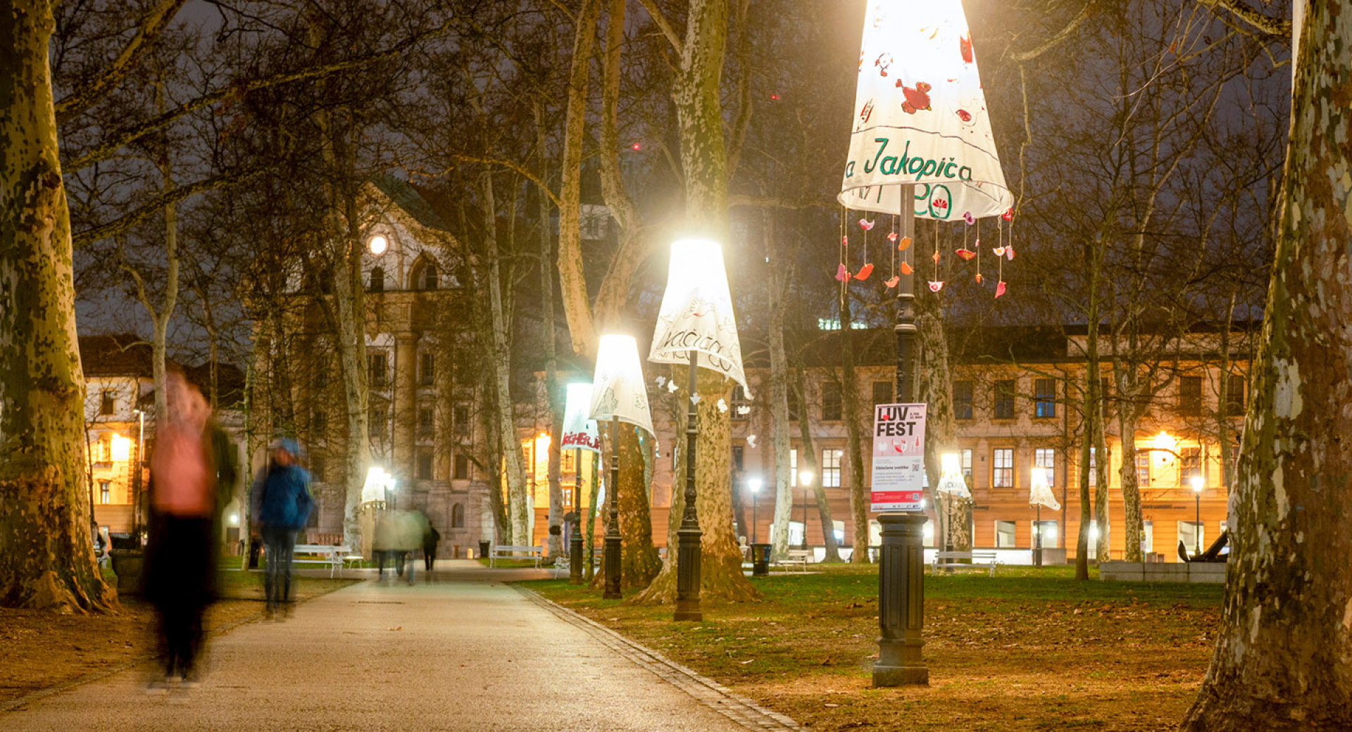 Fotografija tlakovane poti v parku ponoči, obdane z visokimi drevesi in uličnimi svetilkami. Vsaka svetilka je "oblečena" v velik, dekorativen senčnik iz blaga