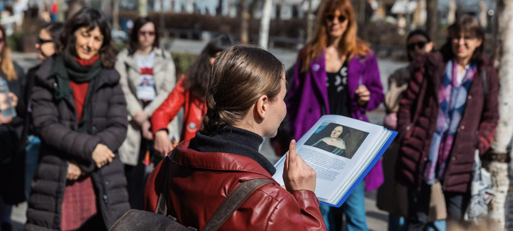 Oseba stoji na prostem in drži odprto knjigo s portretom ženske. V polkrogu stoji skupina več ljudi, ki pozorno posluša.