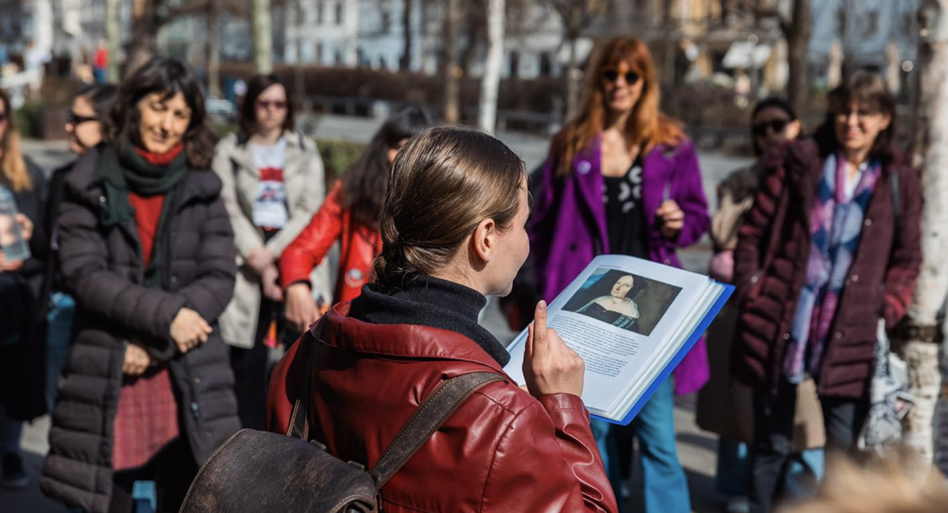 Oseba stoji na prostem in drži odprto knjigo s portretom ženske. V polkrogu stoji skupina več ljudi, ki pozorno posluša.