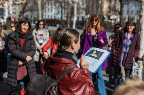 Oseba stoji na prostem in drži odprto knjigo s portretom ženske. V polkrogu stoji skupina več ljudi, ki pozorno posluša.