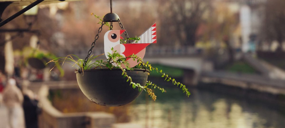 Papirnati pticek v cvetlicni obesanki visi ob zgradbi ob reki.