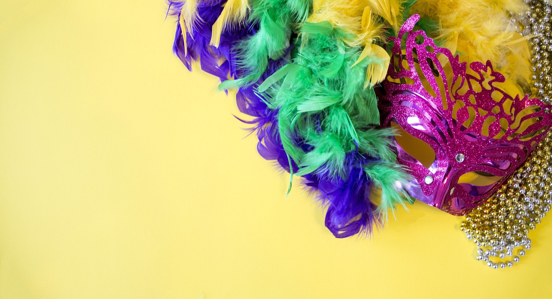 A bright yellow background features a pink glittery mask. Surrounding the mask are colorful feathers ialong with strands of shiny gold beads.