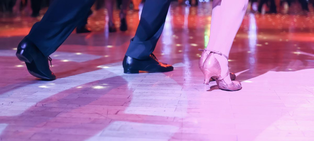 A dancing couple in elegant shoes performs steps on an illuminated dance floor, surrounded by light reflections and other dancers in the background.