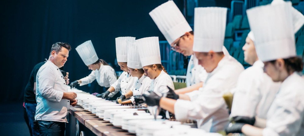 Young chefs prepare dishes while being observed by a master chef.