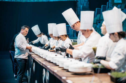 Young chefs prepare dishes while being observed by a master chef.