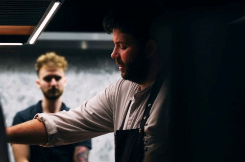 The head chef in a white shirt with a black apron and an observer in a black shirt.
