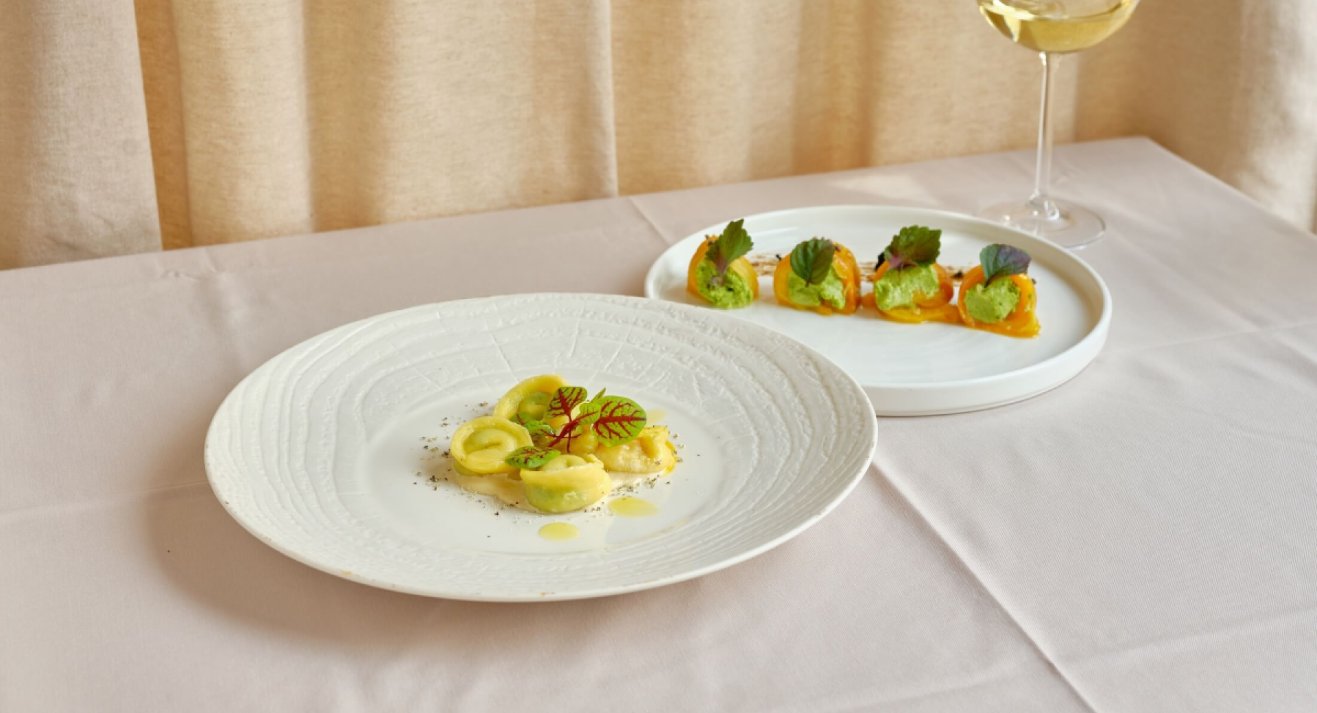A plate features assorted ravioli garnished with decorative greens. Vegetable rolls are served on a plate alongside a glass of white wine.