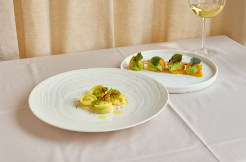 A plate features assorted ravioli garnished with decorative greens. Vegetable rolls are served on a plate alongside a glass of white wine.